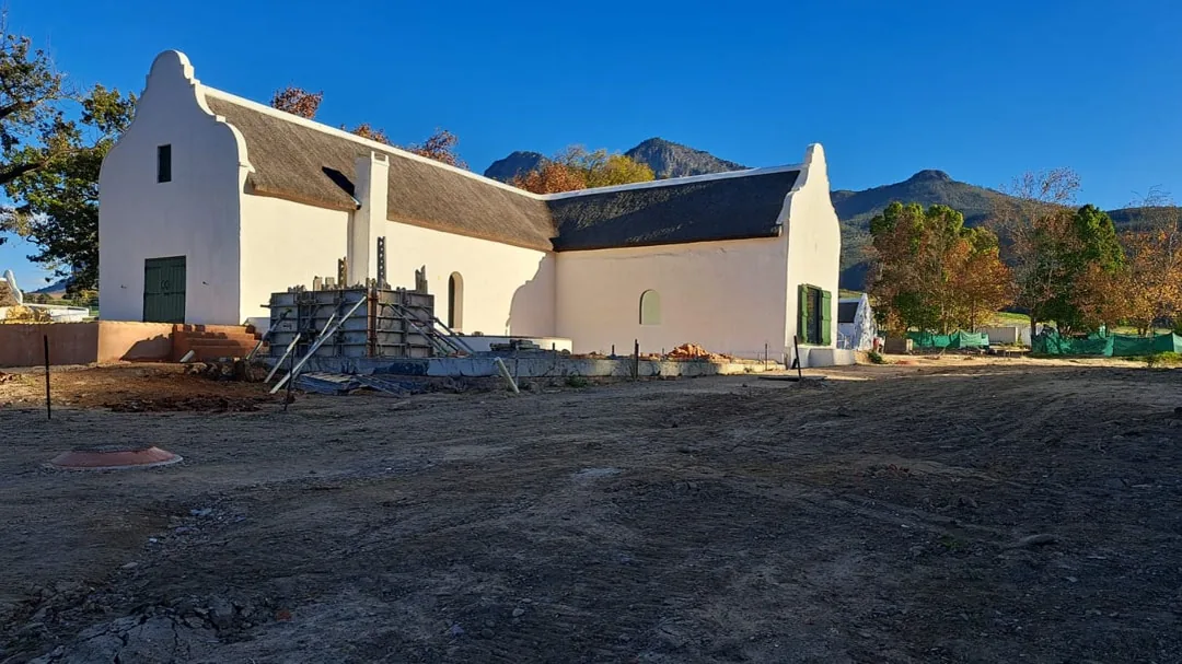 Soetmelksvlei historical farm during restoration by JDV Construction, showing lime-washed Cape Dutch walls, a thatched roof, curved end gables and green shutters against the Winelands mountains
